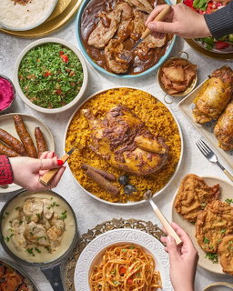 Overhead view of Mediterranean dishes including paella, grilled meats, salads, and bread with hands reaching for food