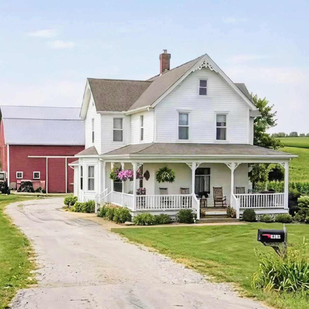 White two-story farmhouse with front porch, red barn, and gravel driveway surrounded by green lawn and trees