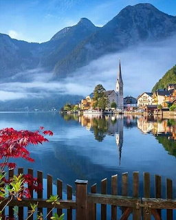 Hallstatt village with church spire reflected in lake, misty mountains in background, autumn foliage in foreground