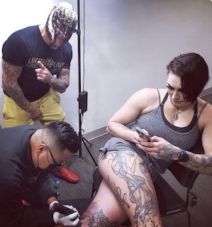 Tattoo artist inking a detailed leg tattoo on a woman sitting in a studio with an observer nearby