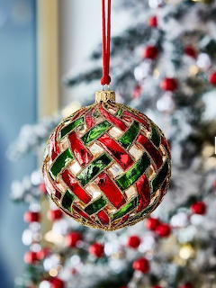 Red and green woven glass Christmas ornament ball hanging by red ribbon against blurred Christmas tree background