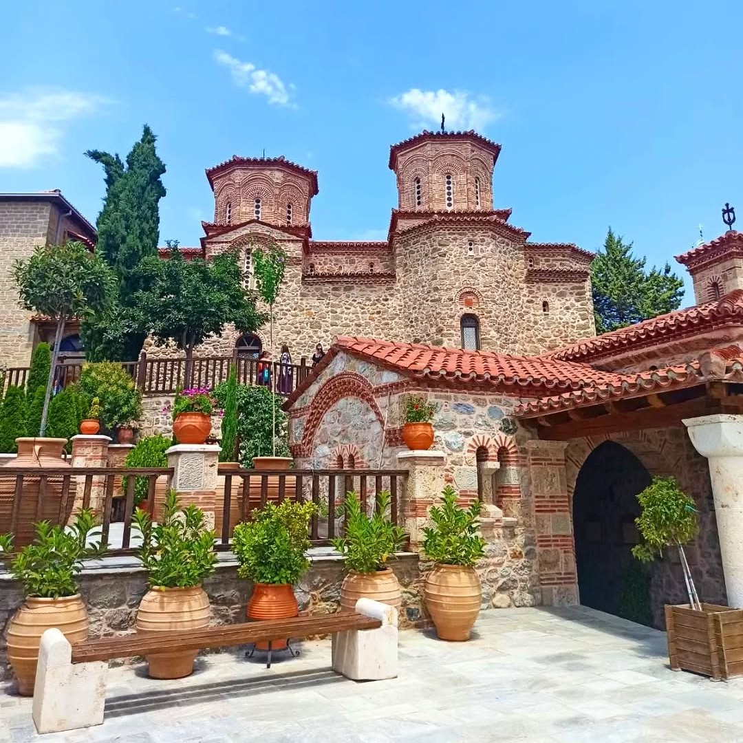 Stone monastery courtyard with tiled roofs, potted plants, and blue sky in Meteora, Greece