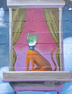 Street art mural showing orange cat in window with pink interior and green curtains on blue wall