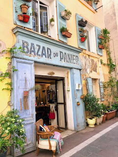 Rustic French boutique with blue weathered storefront, colorful potted plants on balcony, and cobblestone street