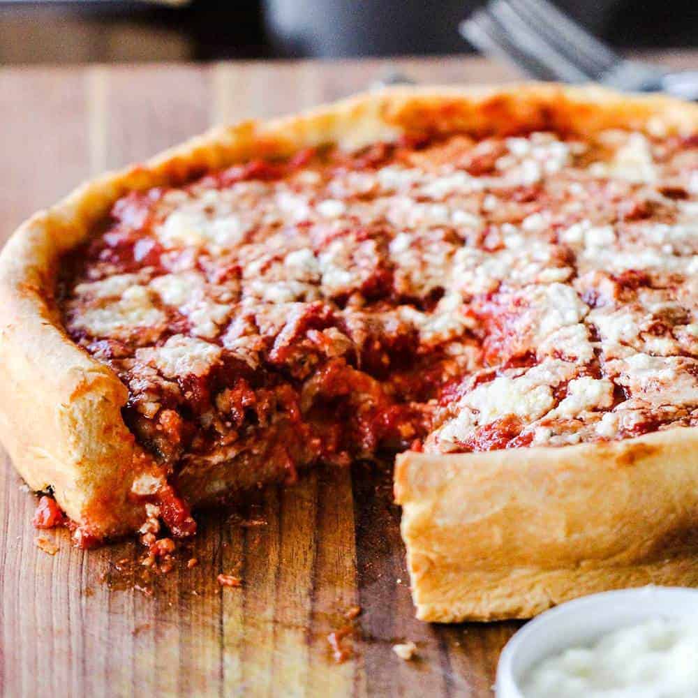 Deep dish pizza with a thick crust on a wooden table next to a small bowl of white dip