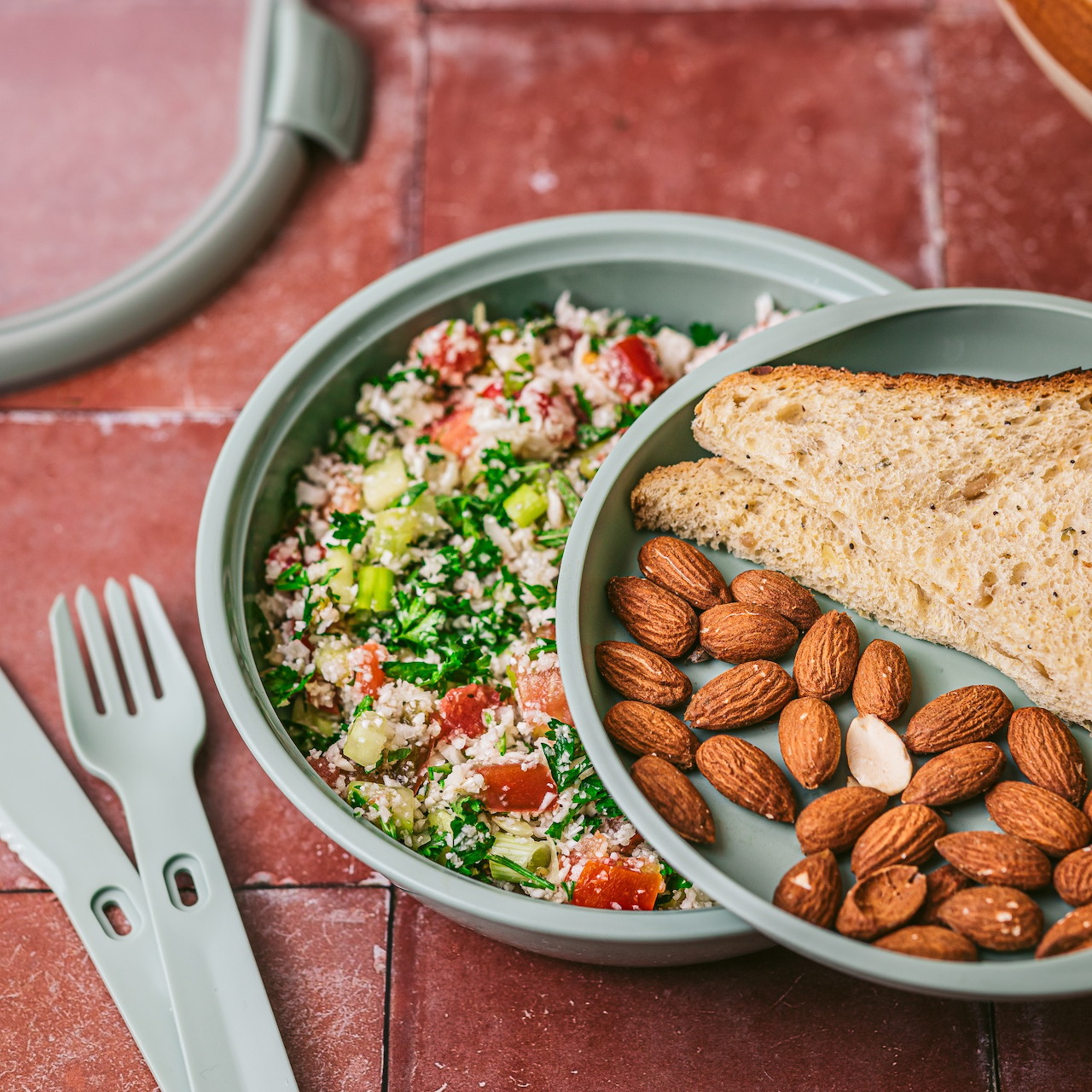 Bowl of grain salad with tomatoes, almonds, and toasted bread beside a fork and spoon on a pink table