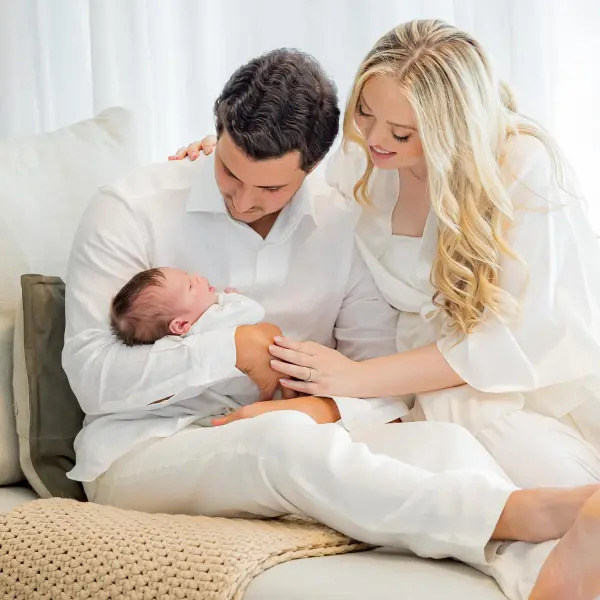 Mother and father in white clothes holding a newborn baby while sitting together indoors
