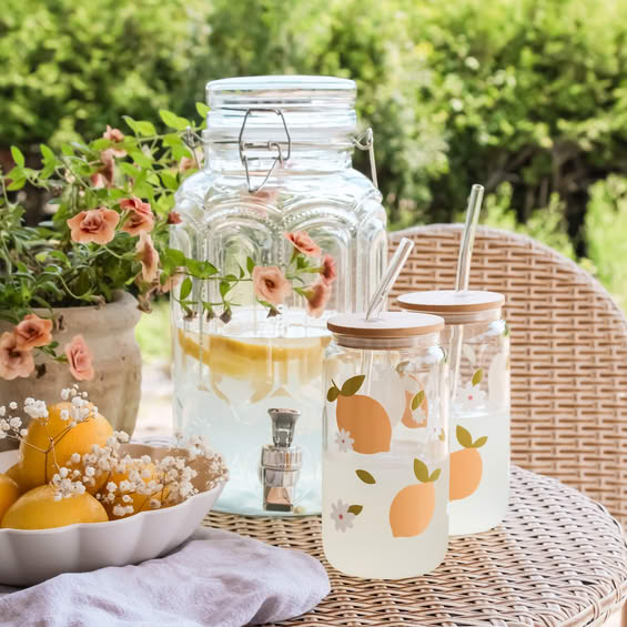 Glass lemonade dispenser with citrus slices on outdoor wicker table with flowers