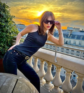 Woman wearing sunglasses leaning on ornate balcony railing at sunset with cityscape behind