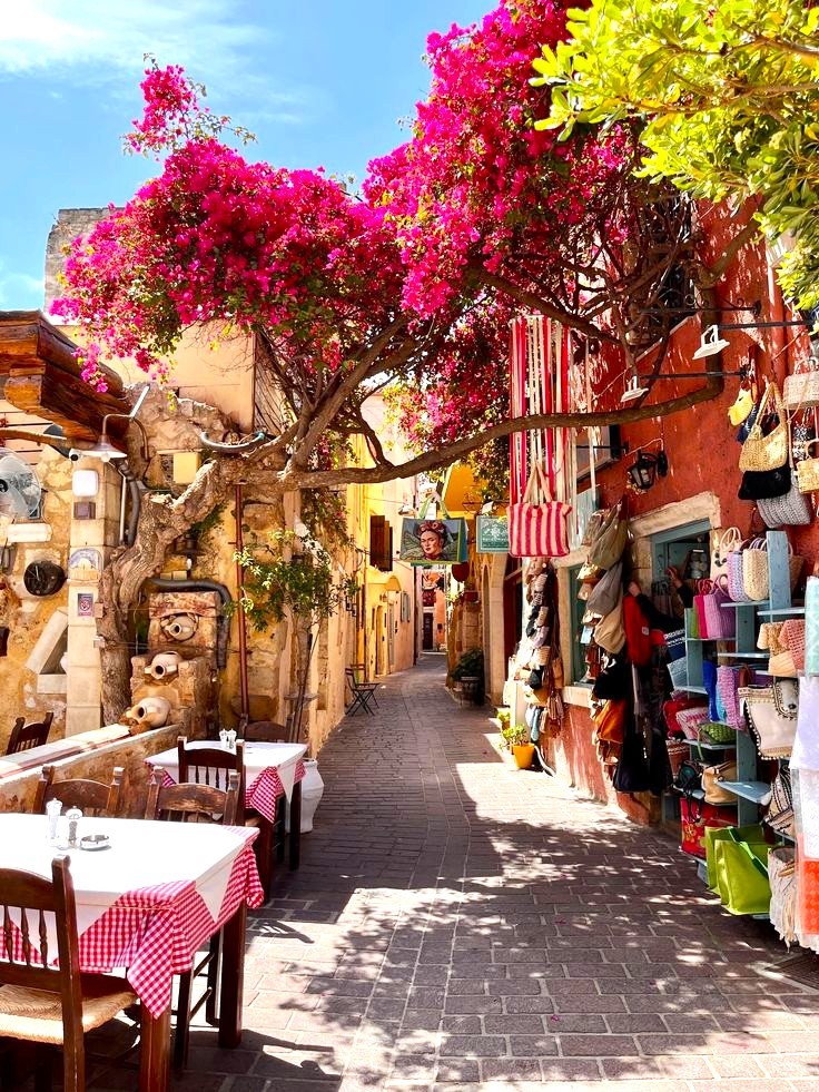 Narrow coblestone street in Crete with pink bougainvillea tree, outdoor cafe tables, and colorful shops