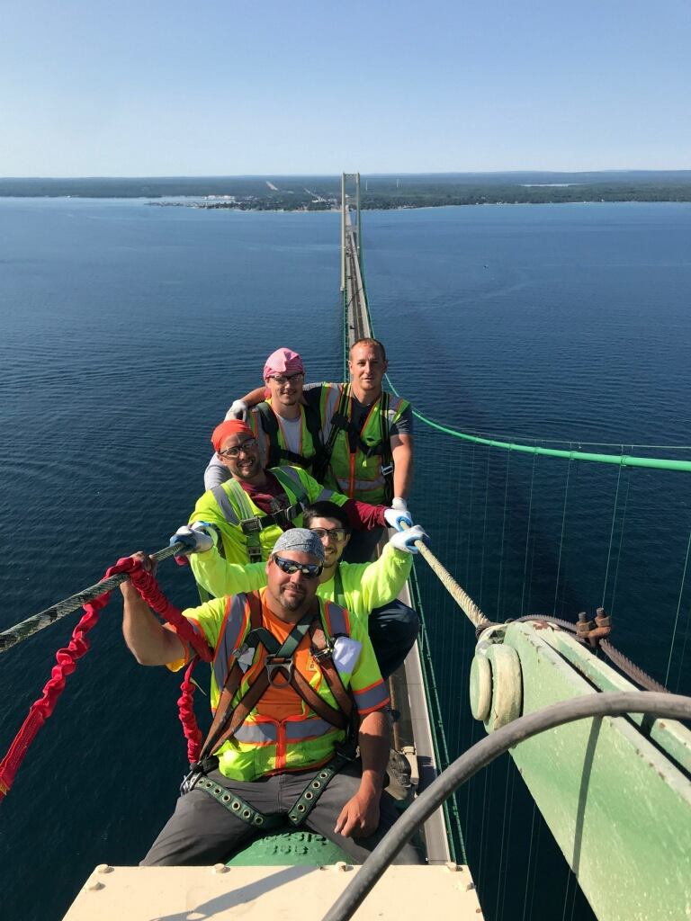 Three workers in safety vests and harnesses on bridge tower above water