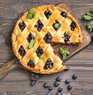 Lattice-topped berry pie with one slice cut, scattered fresh berries and mint on wooden board