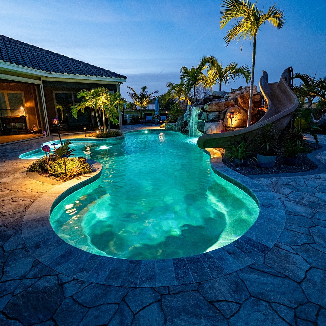 Illuminated turquoise resort pool with palm trees and waterslide at twilight