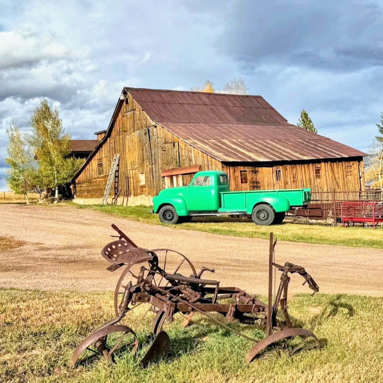 Weathered barn with turquoise vintage truck and rusty farm equipment in rural landscape