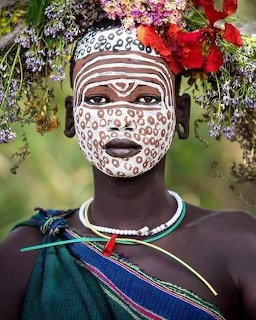Woman with white face paint, flowers on her head, and bead jewelry in a close portrait