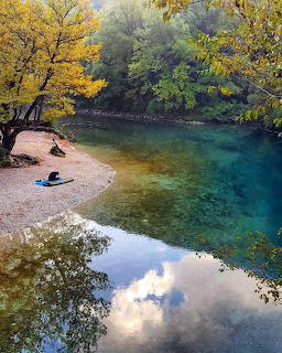 Clear turquoise river bend with autumn trees, pebble shore, and reflections in calm water