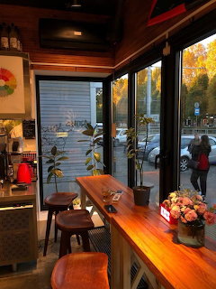 Cozy cafe interior with wooden counter, large windows showing autumn trees, warm ambient lighting