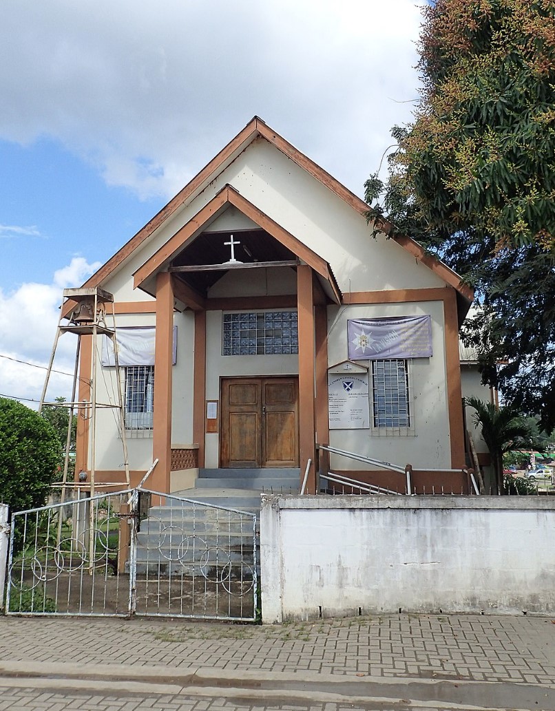 Small church with A-frame roof, cross on facade, wooden door, and metal gate in San Ignacio, Belize