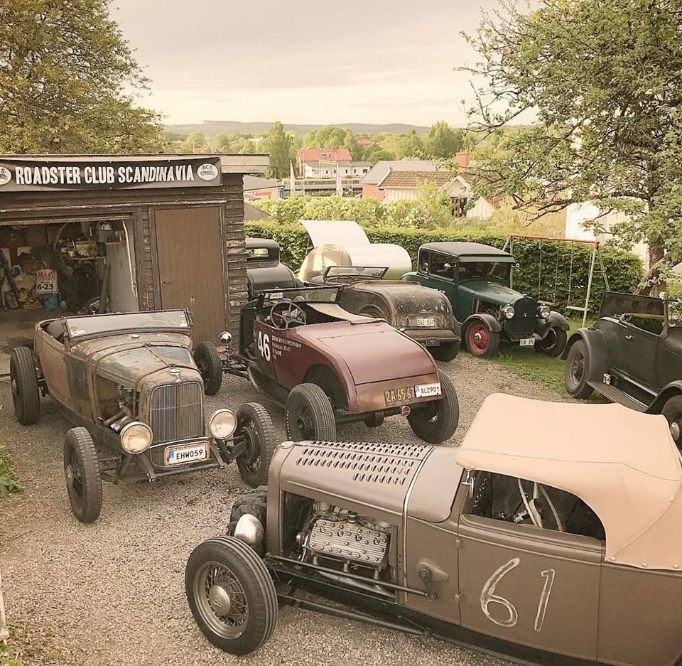 Several vintage hot rods parked in a gravel lot near a garage with trees and houses behind