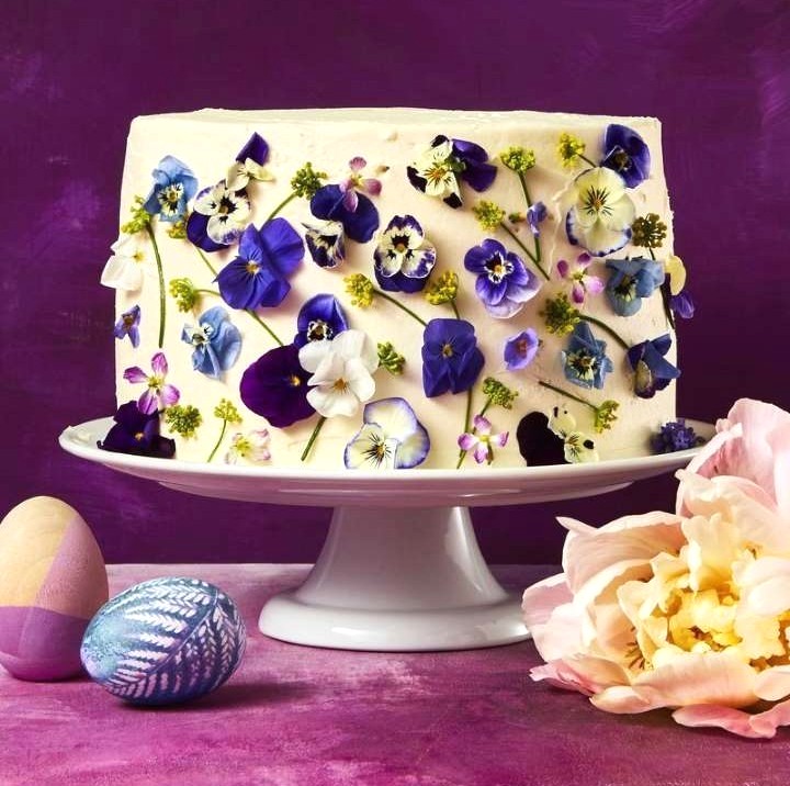 White cake topped with colorful edible flowers on white stand against purple background
