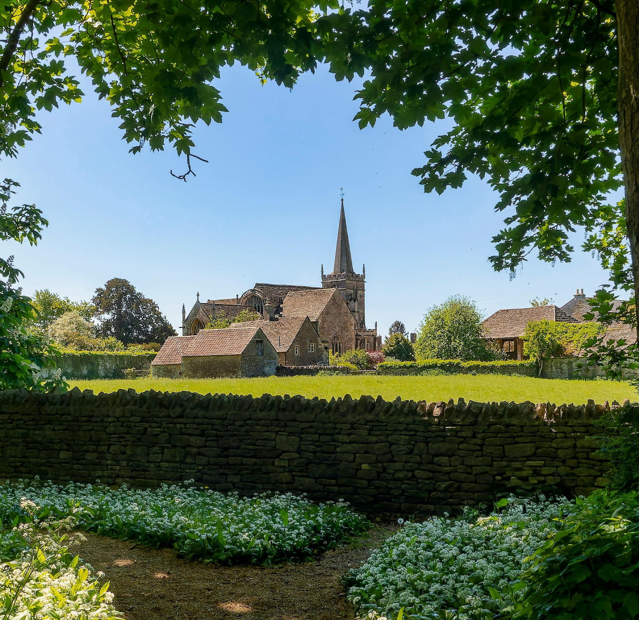 Historic stone church with spire in Cotswold village surrounded by lush green fields and trees