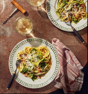 Two plates of pasta with zucchini and cheese on a table with wine glasses and pink napkin