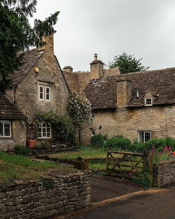 Old stone cottages with tiled roofs, a wooden gate, and garden plants in a village scene.
