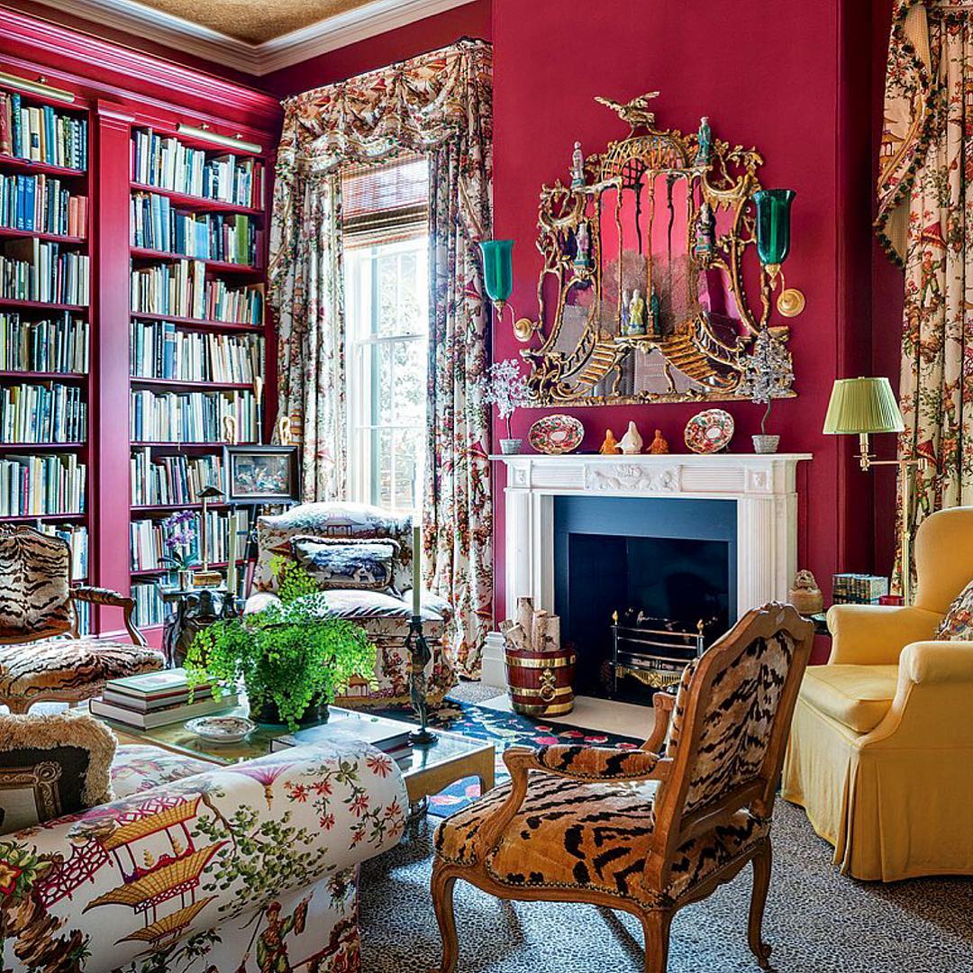 Luxurious library room with red walls, built-in bookshelves, fireplace, floral armchairs, and ornate mirror
