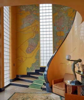 Art deco interior with angular staircase, golden mural walls, white grid panels, and bronze sculpture