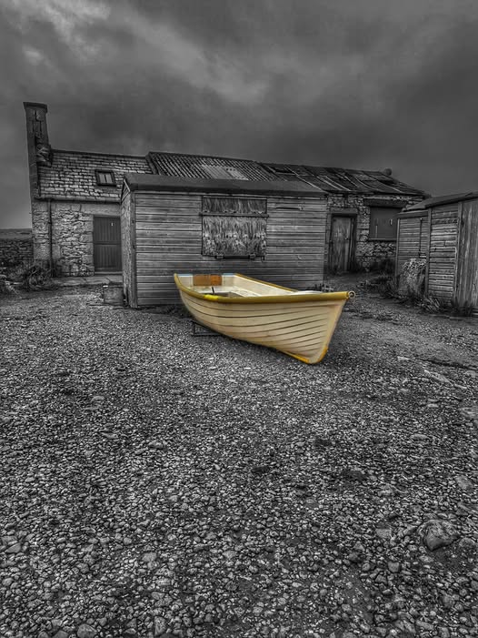 Yellow boat sits on pebble beach in front of old wooden boathouses under dramatic gray sky