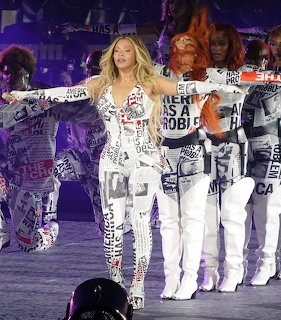Beyoncé performing on stage in a white text-printed outfit with backup dancers behind her
