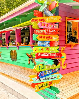 Colorful hand-painted wooden direction signs to world cities in front of a bright pink tropical hut