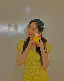 Young woman in a yellow dress holding a small colorful item beside her face indoors