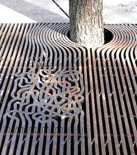 Decorative metal tree grate with wavy parallel lines and ornate cutout pattern around a tree trunk