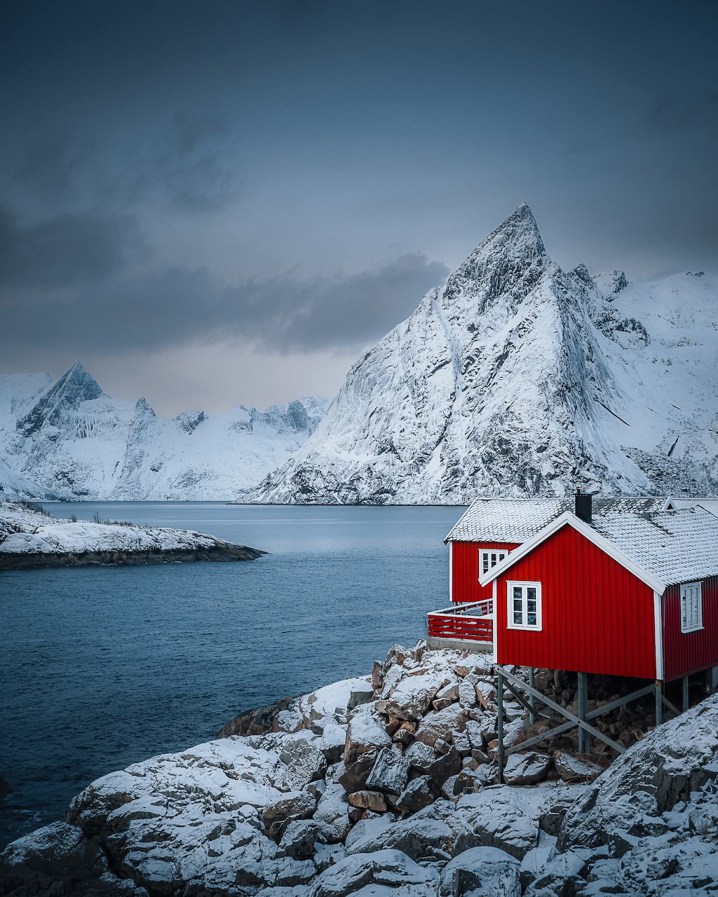 Red Norwegian cabin on snowy rocks beside a fjord with snow-covered mountain peaks in winter
