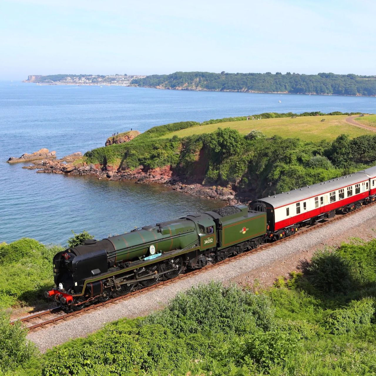 Green steam train with carriages on a coastal railway beside the sea and grassy cliffs