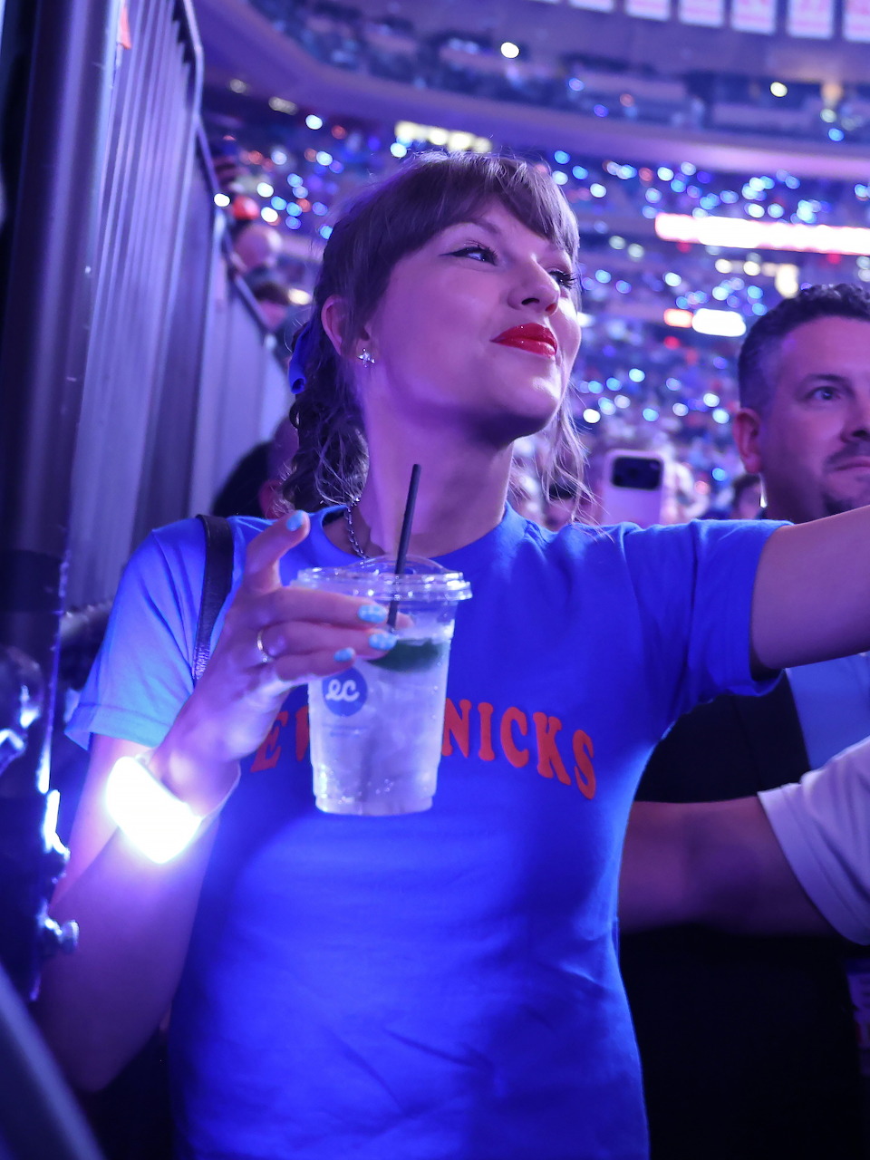 Woman in a blue Knicks shirt holding a drink at a brightly lit arena event