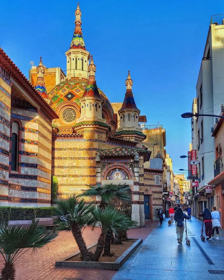 Colorful mosaic-decorated church of Sant Romà in Lloret de Mar with palm trees and pedestrian street at dusk