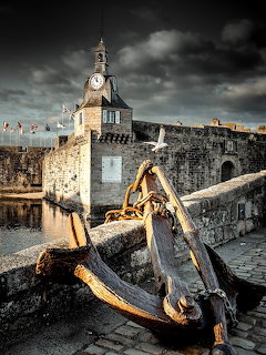 Rusty anchor on stone quay with walled medieval town and clock tower under dramatic sky