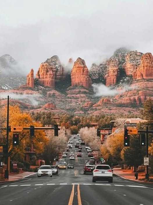 Road leading into Sedona with cars, autumn trees, and misty red rock cliffs in the background