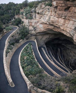 Curved paved pathway leading into massive cave opening with striped rock walls and greenery