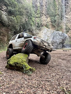 Muddy Mitsubishi Pajero SUV perched on a mossy rock in a forest clearing
