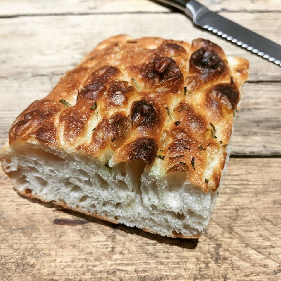 Slice of focaccia bread with golden crust and herbs on wooden board, showing soft interior crumb