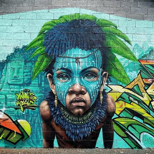 Street mural of an Indigenous woman with turquoise face paint and a green feathered headdress.
