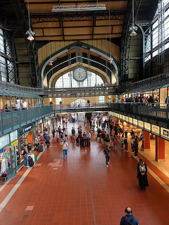 Interior of Hamburg Hauptbahnhof station hall with a central clock, shops, and people walking below
