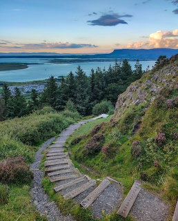 Stone steps descending a green hillside overlooking a bay with mountains and sunset sky