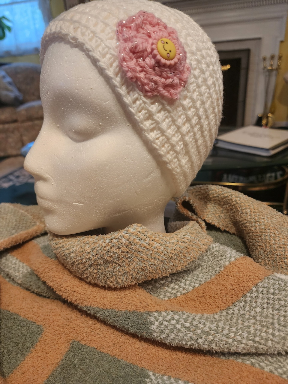 White mannequin head wearing a cream knit hat with a pink flower and a striped scarf indoors
