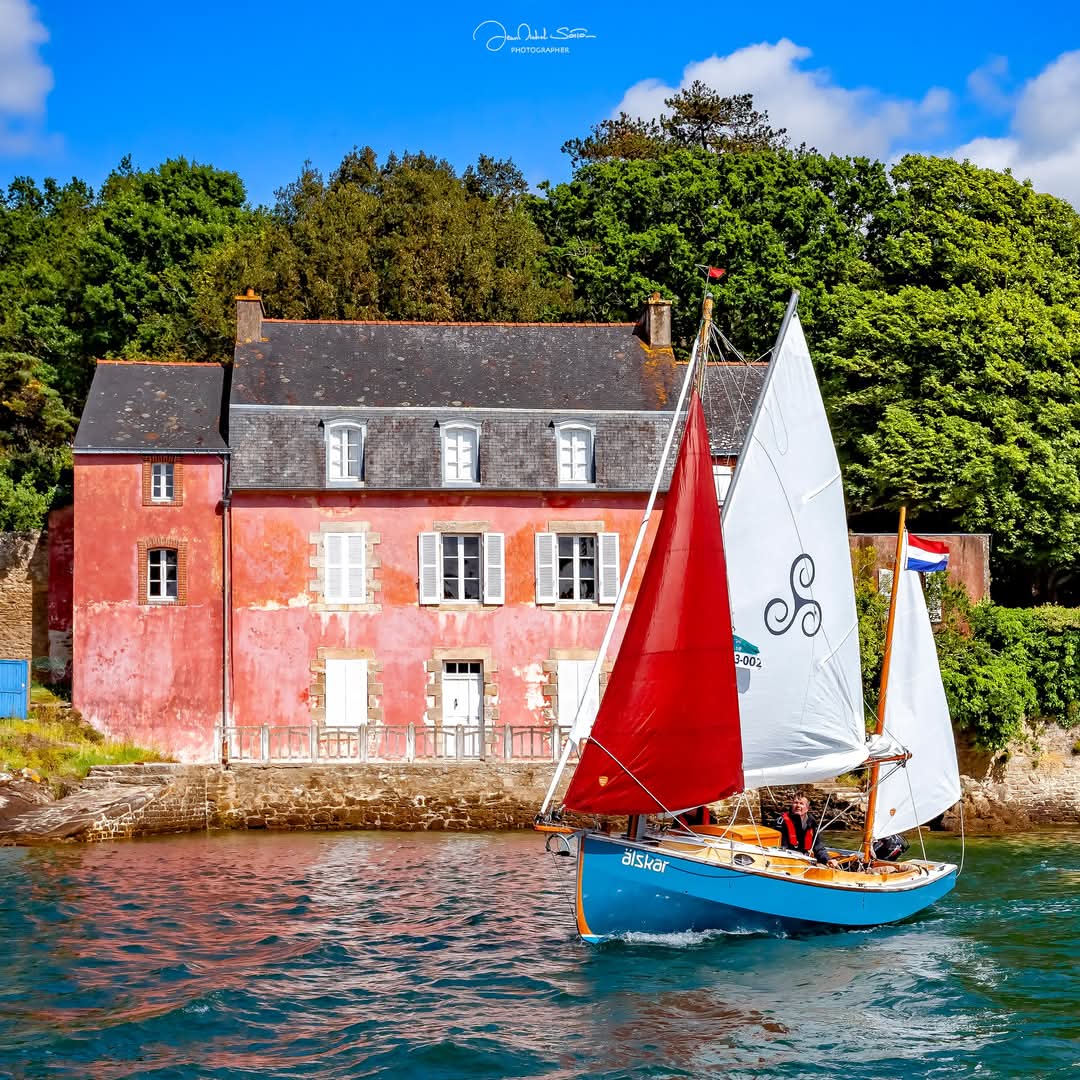 Sailboat with red and white sails passing a pink house by the water