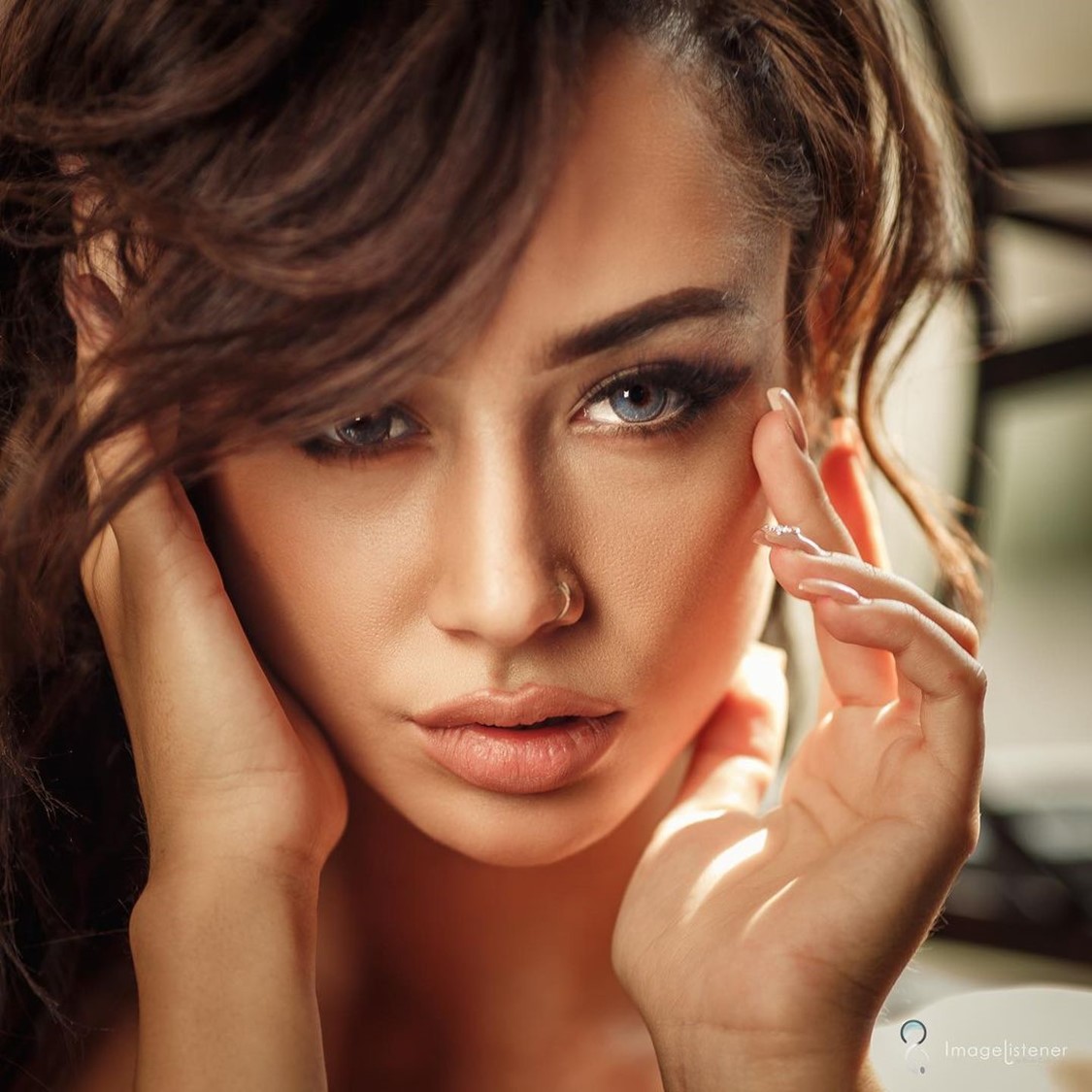 Close-up portrait of a woman with brown wavy hair, smoky makeup, and a nose ring