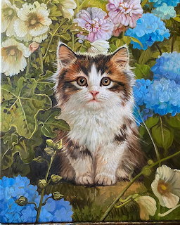 Fluffy tabby kitten sitting in garden surrounded by colorful flowers including blue hydrangeas and pink blooms